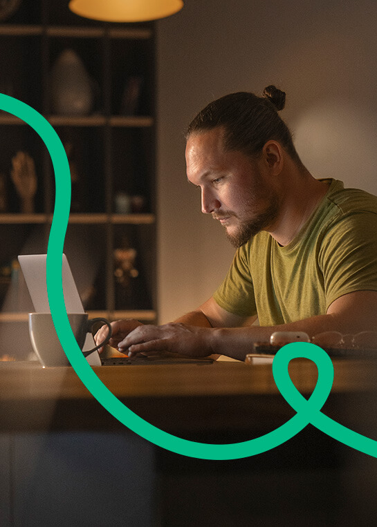 Fiatal férfi zöld pólóban, összpontosítva dolgozik egy laptop előtt egy hangulatos, félhomályos szobában. Egy csésze kávé van mellette az asztalon. A kompozíciót egy dinamikus, zöld, hullámos vonal egészíti ki.